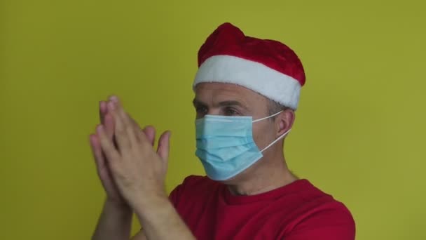 Een man met een kerstmuts met een medisch masker applaudisseert en gebaart met zijn handen. — Stockvideo