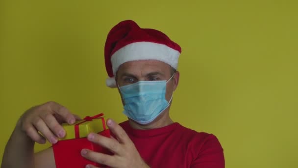 Een man met een kerstmuts en masker houdt een geschenkdoos in zijn hand.. — Stockvideo