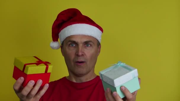 Un atractivo hombre de 40-50 años en un sombrero de Santa felizmente ofrece cajas de regalo. — Vídeo de stock