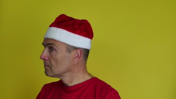 Человек в шляпе Санты в красной футболке на жёлтом фоне поворачивает голову налево и направо. Мужчина смотрит вверх. — стоковое видео