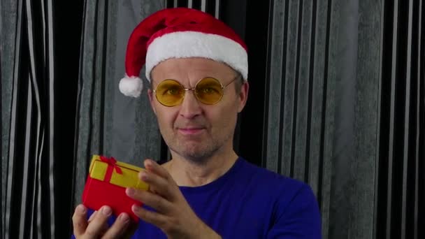 Человек в шляпе Санта Клауса смотрит на него и трясет подарочной коробкой.. — стоковое видео