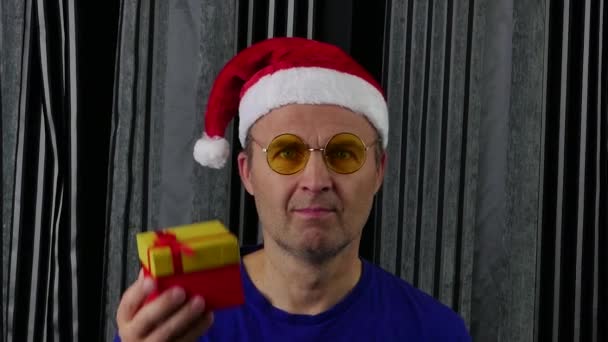 サンタの帽子と眼鏡をかけたハンサムな男がプレゼント箱を持っている. — ストック動画