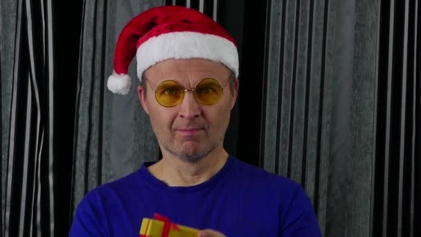 Un bell'uomo con un cappello da Babbo Natale si toglie gli occhiali e guarda la confezione regalo. — Video Stock