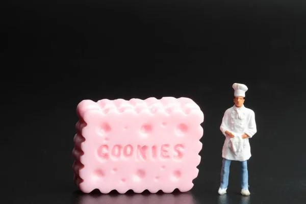Miniature Figurine Cook Huge Biscuit Black Background — Zdjęcie stockowe