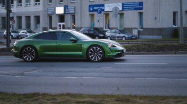 Riga, Letonya - 10 Nisan 2021: yeşil Porsche Taycan soğuk bir bahar akşamında şehir binalarının arka planına doğru hızla sürüyor