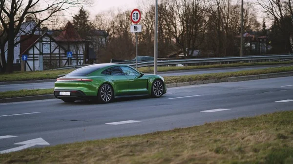Riga, Letonya - 10 Nisan 2021: yeşil Porsche Taycan soğuk bir bahar akşamı şehir parkının arka planına doğru sürüyor