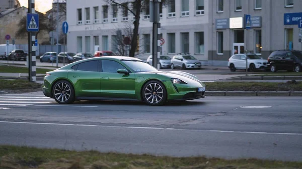 Riga, Letonya - 10 Nisan 2021: yeşil Porsche Taycan soğuk bir bahar akşamında şehir binalarının arka planına doğru hızla sürüyor