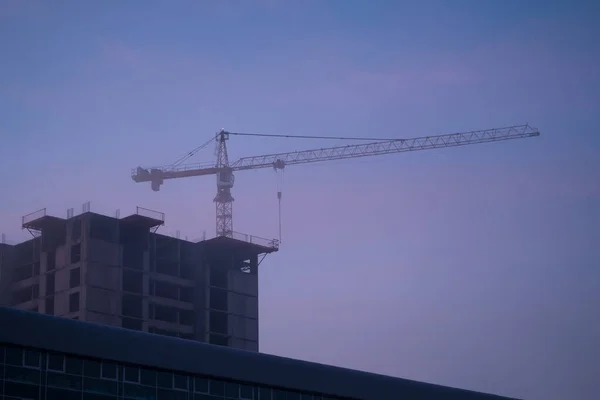 Güneşin doğduğu sisli bir sonbahar sabahında inşaat vincinin silueti ve bitmemiş konut binası. Ev inşaatı, apartman bloğu
