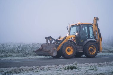 Riga, Latvia-October 6, 2021: Yellow JCB construction tractor is driving on the foggy road during the cold autumn morning