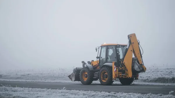 Yellow construction tractor is driving on the foggy road during the cold autumn morning