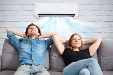 Happy couple sitting on sofa under air conditioner at home