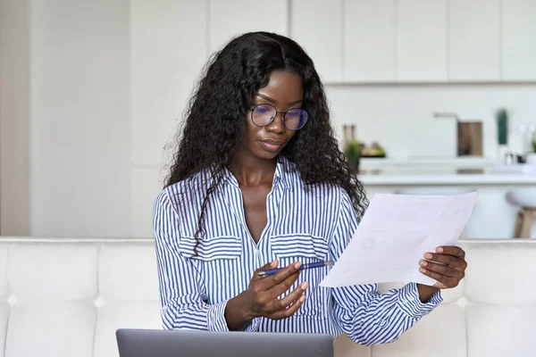 Young black woman holding paper working on laptop at home office.