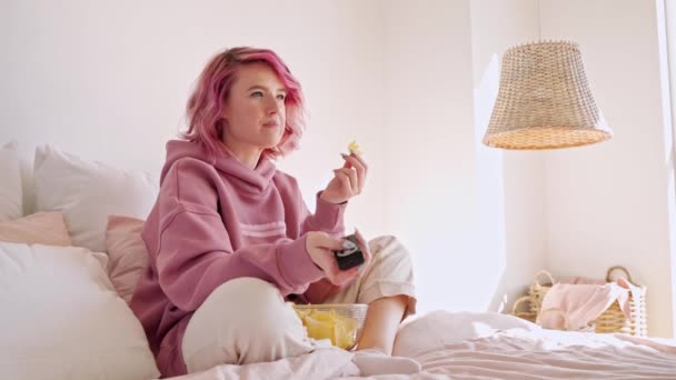 坐在床上 拿着遥控器 吃着薯片吃着粉红头发电视系列电影的少女 — 图库视频影像
