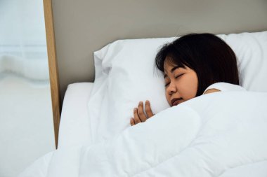 Asyalı kadın yatak odasında beyaz yatakta tatlı rüyalar görür ve evde iyi dinlenir.
