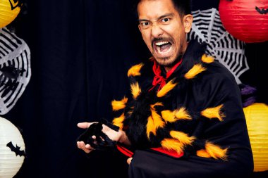 Asyalı genç büyücü Cadılar Bayramı erkek portresi ve göğsünde fotokopi alanıyla yakın plan siyah örümcek. Siyah arka planda Cadılar Bayramı kostümünde moda sanatının tasarımına sahip büyücü makyajı çekici bir model