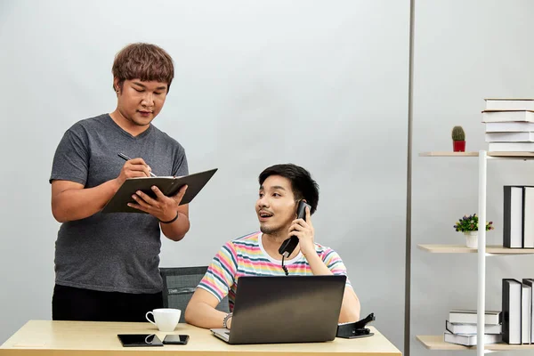 Asian Gay couple at office using internet and laptop shopping online purchase. Fat guy standing writing memo from friend conversation by telephone