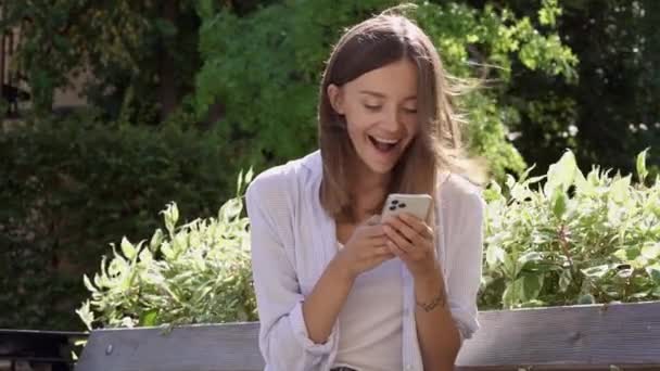 幸せな若い女性がベンチに座って電話を見ている 幸せな反応と良いニュースの感情 — ストック動画