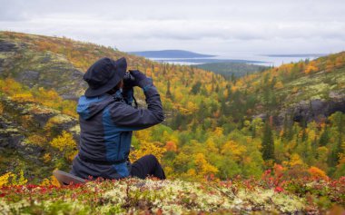Dağın yamacında şapkalı bir kadın, sonbaharda bir gölün ve dağların arka planına dürbünle bakar. Arkadan bak. fotoğraf