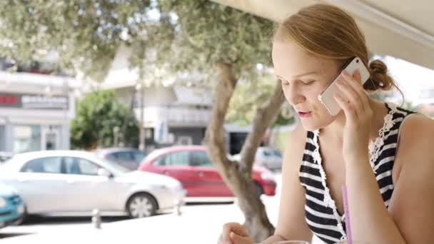 Kobieta siedzi na miejskich kawiarni obok ulicy rozmawia przez telefon komórkowy — Wideo stockowe