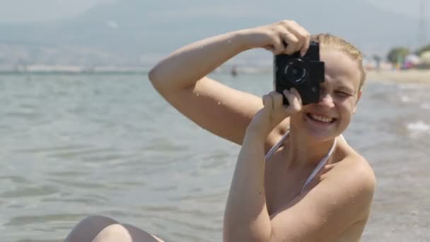Mujer sonriente en bikini sentada en la playa al borde del agua tomando una fotografía — Vídeo de stock