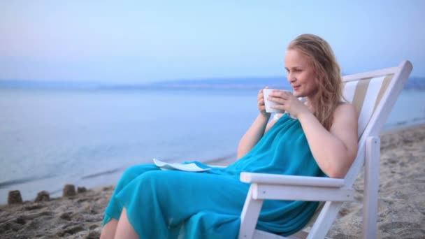 Женщина, наслаждающаяся чашечкой чая на берегу моря, расслабляясь на шезлонге — стоковое видео