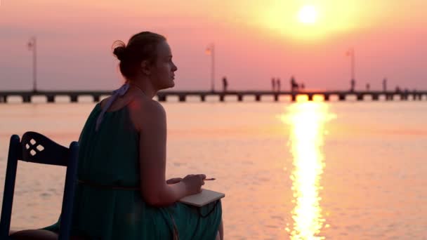 若い女性が座って日没時に海岸で彼女の日記に何かを書く — ストック動画