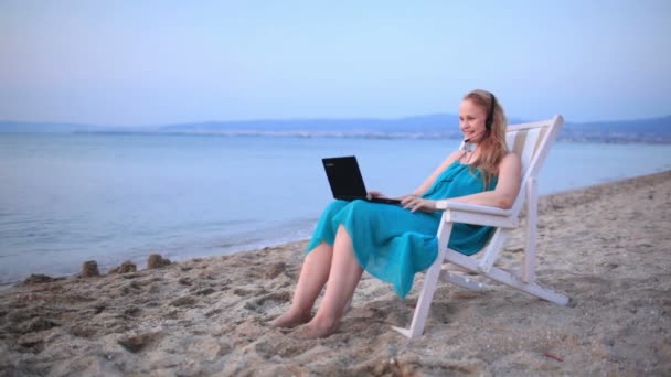 Mulher relaxante na praia com seu computador portátil e vestindo um fone de ouvido conversando com seu amigo com skype — Vídeo de Stock