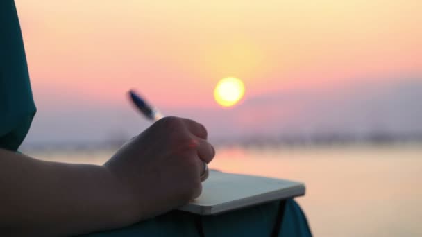 Κλείνω πάνω θέα του χεριού του μια γυναίκα που γράφει στο ημερολόγιό της στο ηλιοβασίλεμα — Αρχείο Βίντεο