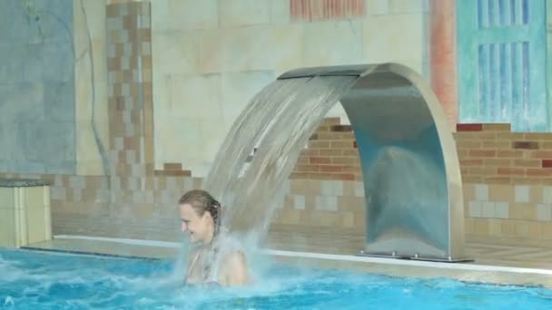 Mädchen spielt in einem Schwimmbad, das unter einem Wasserstrahl aus einem gebogenen Wasserspiel am Pool steht — Stockvideo