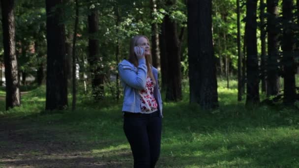 Junge Frau im Park geht langsam mit Blatt in der Hand und telefoniert — Stockvideo