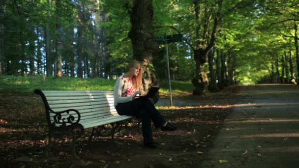 Mujer joven sentada en el banco en el parque y usando su computadora portátil — Vídeo de stock
