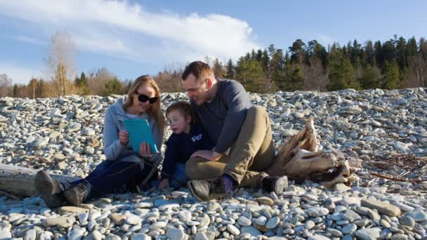 Семья из трех человек сидит на каменистом берегу — стоковое видео