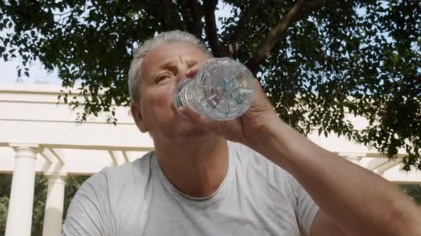 A sporty elderly caucasian man drinks water after fitness classes — Stock Video