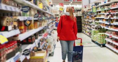 Koruyucu maskeli kadın süpermarketten dolu bir arabayla geçiyor ve başka bir şeye ihtiyacı var mı diye etrafa bakıyor.