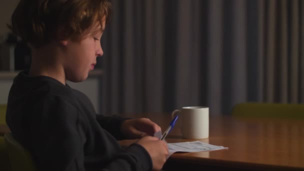男孩在晚上做作业 他拿起文件填好了答案 他嘴里衔着笔 在想一些困难的任务 — 图库视频影像