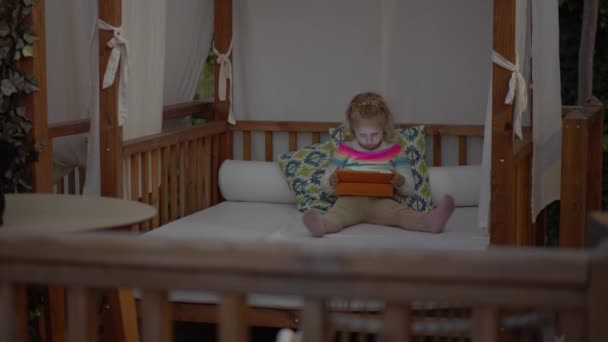 一个手里拿着平板电脑的高加索小女孩在玩电脑游戏 她坐在一个舒适的沙发上 沙发在街道上 盖着白色的桌布 — 图库视频影像