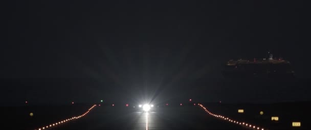 飞机夜间在灯光明亮的跑道上起飞 前照在过往的游轮上 — 图库视频影像