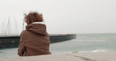 Back view of a boy in warm jacket sitting alone on the coast and watching the ocean waves crushing the breakwater with sailboats parking