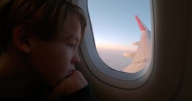 Close-up shot of a boy looking through the plane window being tired and bored with a long flight, aircraft wing outside