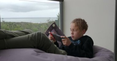 Teenage boy relaxing at home with video games. Child lying on beanbag bag by the window with scenic landscape and spending home leisure with handheld game console