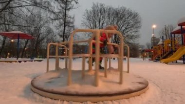 Little girl playing alone on the playground near the house. Winter fun on merry-go-round in the evening