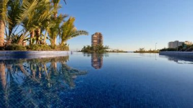 Cinemagraph - Outside swimming pool sided with green plants with view to the clear blue sly and few buildings, water slightly rippling. Resort town scene