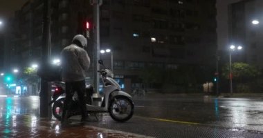 VALENCIA, İSPAN - 13 Temmuz 2022: yağmurlu bir gece. Yağmurluklu bir adam park etmiş scooterına yaklaşıyor, telefonu tamir ediyor ve yola çıkıyor.