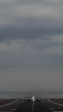 Dikey video - bir yolcu uçağı hızını arttırıyor, su kenarından başlayıp kalkış yapan bir pistte hızlanırken arka plandaki diğer uçak iniş için yaklaşıyor.