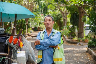 Maske takan mutlu Asyalı bir sokak temizleyicisinin portresi işe gitmeden önce eski bir çöp arabasının yanında kollarını kavuşturuyor..