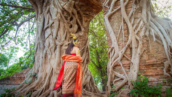 タイの伝統的なドレスのアジアの女性は大きな根の木のカバー壁と古いレンガ造りの門に歩く — ストック写真