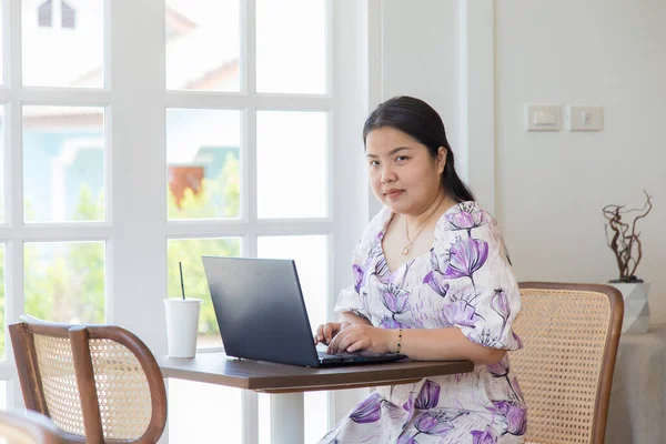 Beyaz desenli elbiseli Asyalı bir kadın kahve dükkanındaki bir masada dizüstü bilgisayarla çalışıyor..