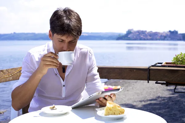 Young handsome man having breakfast on lake reading tablet — Stock Photo, Image