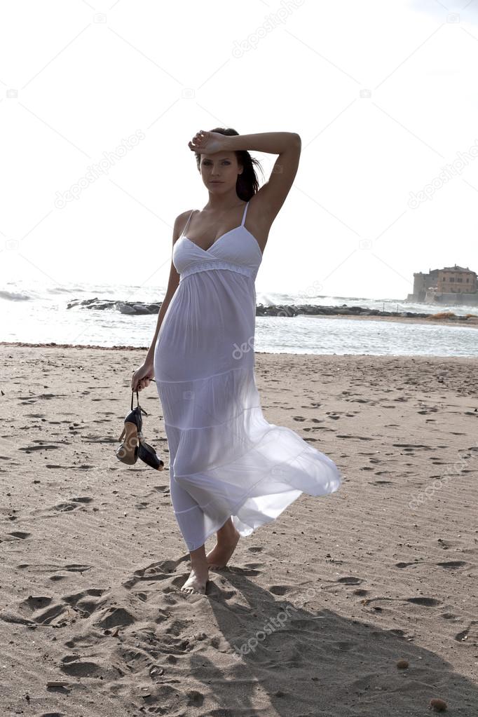 Woman with white dress in front of ocean with shoes in hand