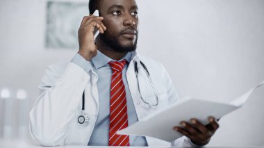 Afro-Amerikan doktor elinde bulanık dosya tutarken cep telefonuyla konuşuyor. 